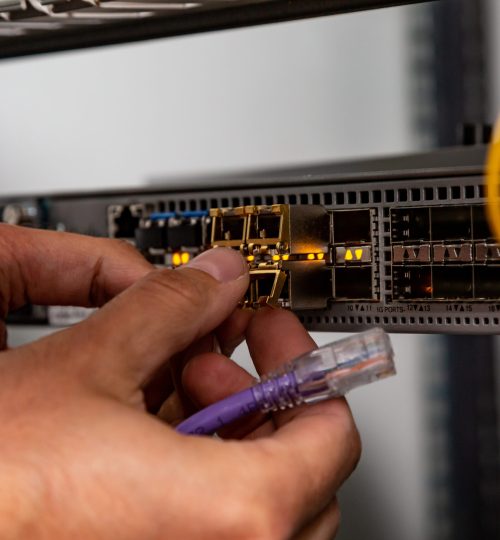 The electrician inserts the SFP module into the router and holds the patch cord. Hands in the frame. Small form-factor pluggable transceiver. Horizontal orientation. High quality photo.